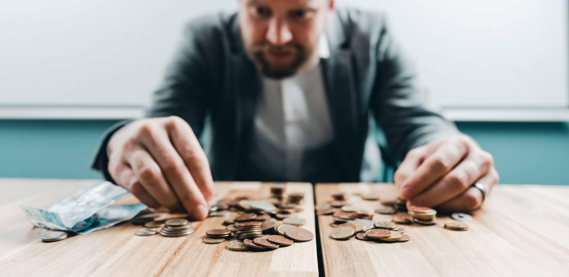 Kuvassa etualalla on kolikoita pinoissa ja kasoissa pöydällä ja taustalla oleva mies laskee rahoja sormillaan.