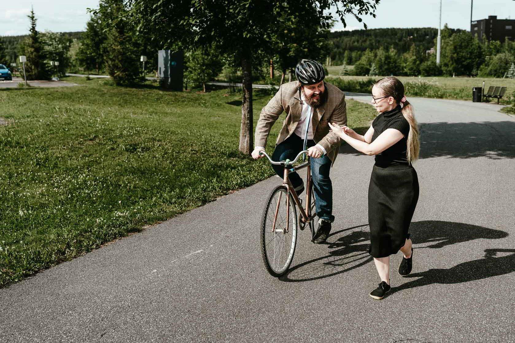 Kuvassa mies polkee pyörällä ja nainen juoksee hänen vierellään näyttäen samalla puhelimesta jotain miehelle.